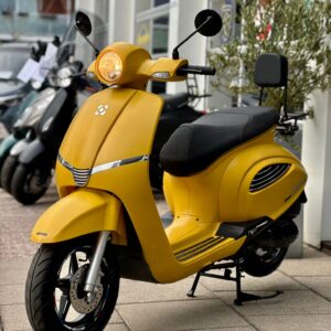 Felgele scooter met brandende koplamp, geparkeerd voor een scootershowroom op de stoep.
