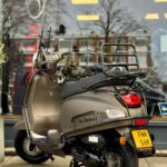 Matbronzen scooter met windscherm en bagagedrager, geparkeerd op standaard voor een winkelraam.