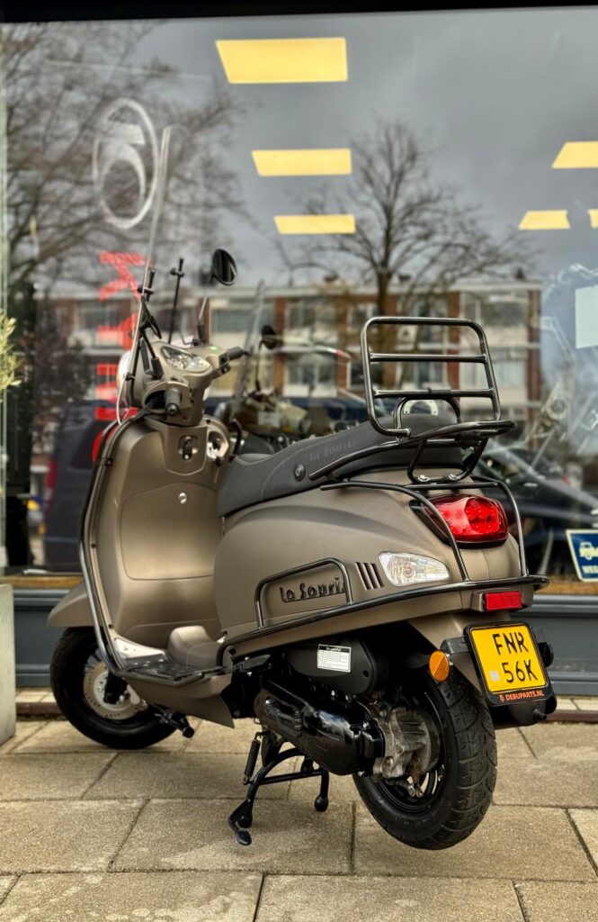 Matbronzen scooter met windscherm en bagagedrager, geparkeerd op standaard voor een winkelraam.