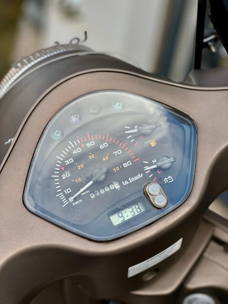 Close-up van het dashboard van een scooter met snelheidsmeter, brandstofmeter en digitaal klokje 9:38.