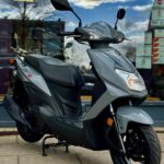 Grijze scooter met moderne lijnen op de standaard, voor een winkelruit, vooraanzicht vanaf rechts.