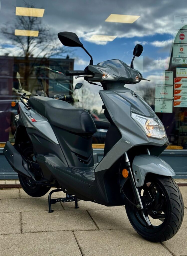 Grijze scooter met moderne lijnen op de standaard, voor een winkelruit, vooraanzicht vanaf rechts.