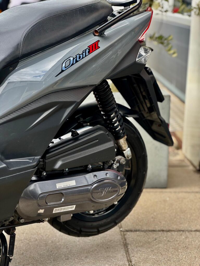 Close-up van de achterzijde van een grijze SYM Orbit III scooter met veer en motorblok.