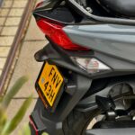 Detail van de achterzijde van een grijze scooter met rode achterlichten en geel kenteken, geparkeerd op de stoep.