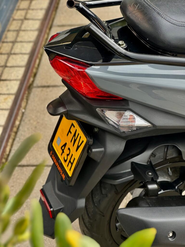 Detail van de achterzijde van een grijze scooter met rode achterlichten en geel kenteken, geparkeerd op de stoep.