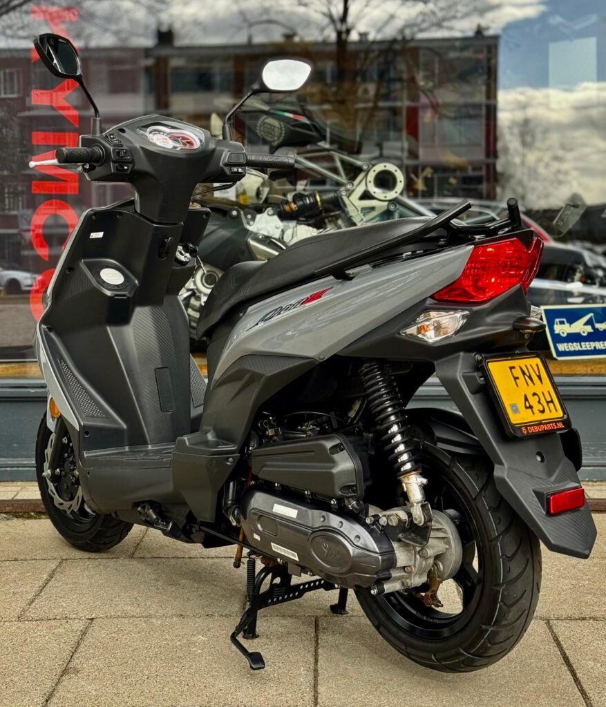 Grijze scooter op middenbok voor een winkelruit, met zichtbaar Nederlands kenteken.