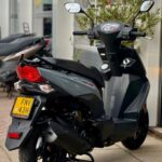 Grijze scooter met spiegels geparkeerd voor een showroom, achteraanzicht op middenstandaard.