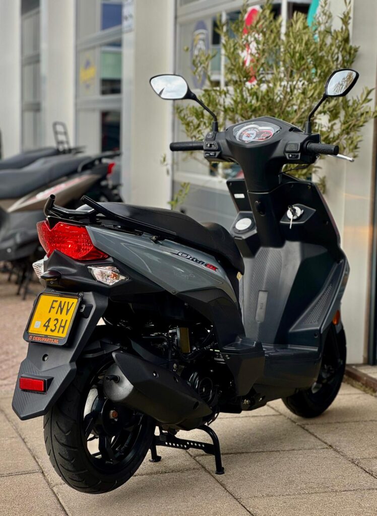 Grijze scooter met spiegels geparkeerd voor een showroom, achteraanzicht op middenstandaard.