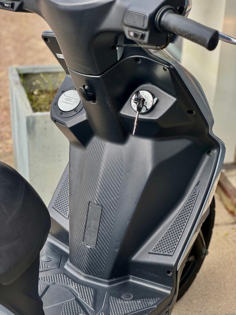 Close-up van het stuur en de voetenplank van een zwarte scooter met sleutel in het contact.