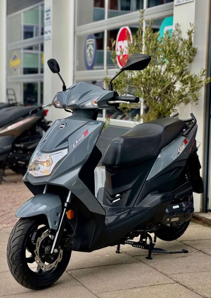 Grijze scooter op de standaard met schijfrem, geparkeerd voor een showroom met logo's.