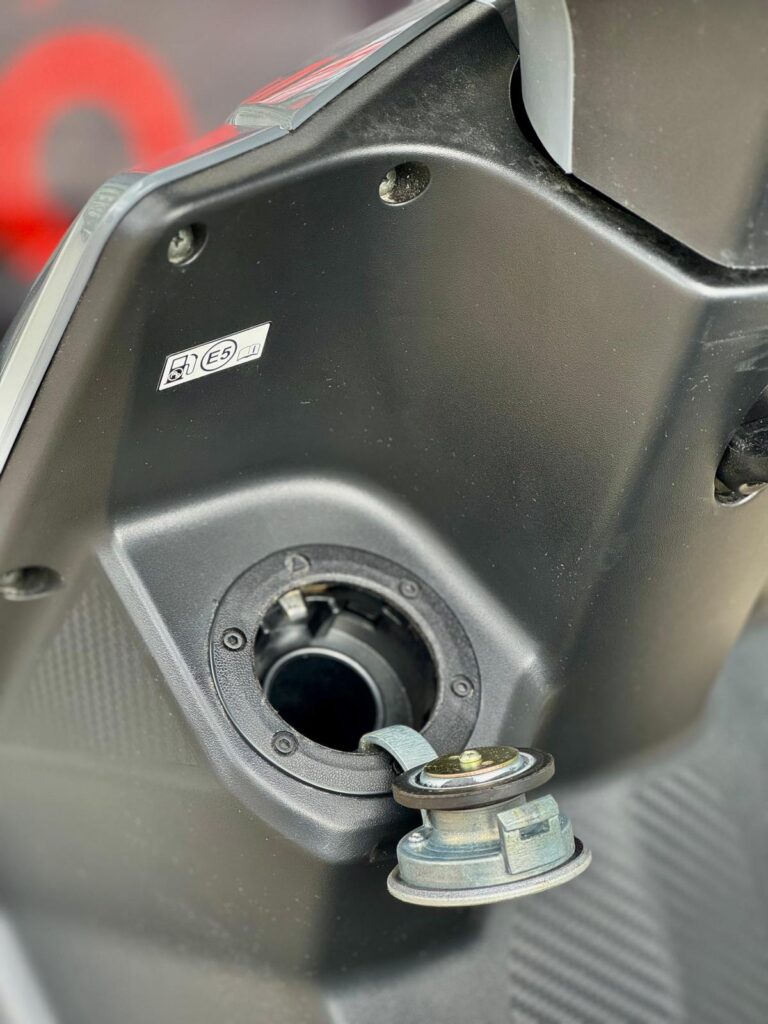 Close-up van open brandstofklep en tankdop van scooter met E5-sticker.