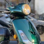 Close-up van een groene scooter met brandende koplamp en oranje reflector, geparkeerd buiten.