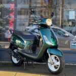 Groene retro-scooter met brandende koplamp op de standaard, geparkeerd voor een winkelraam.
