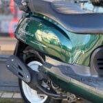 Close-up van de achterzijde van een donkergroene scooter met zwart zadel, wiel en uitlaat.