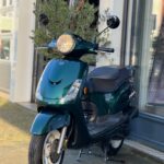 Donkergroene scooter met brandende ronde koplamp, op standaard voor een winkelpui.