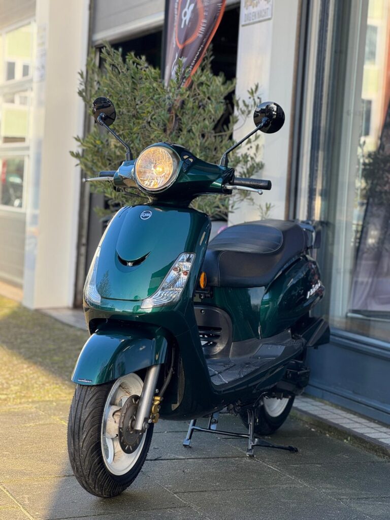 Donkergroene scooter met brandende ronde koplamp, op standaard voor een winkelpui.