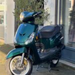 Donkergroene scooter met ronde koplamp geparkeerd op de stoep voor een etalage.