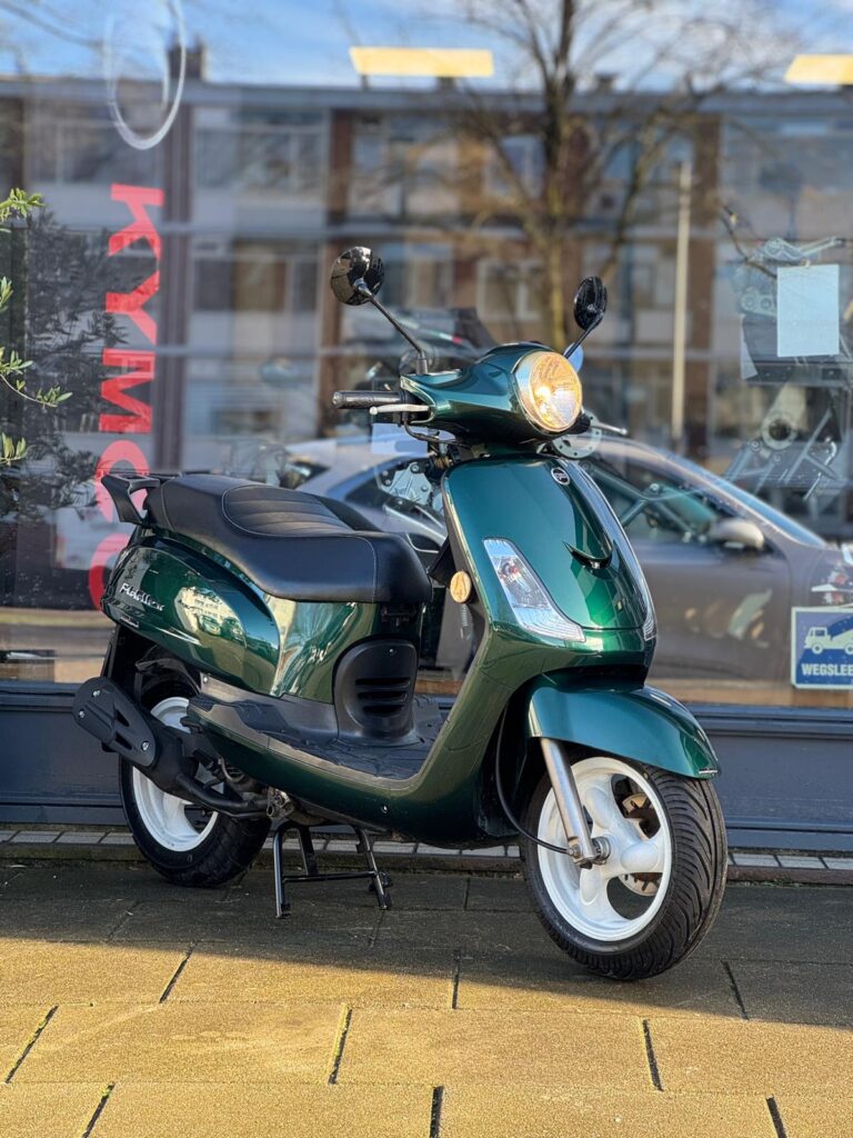 Groene retro-scooter met brandende koplamp op de standaard, geparkeerd voor een winkelraam.
