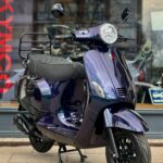 Paars metallic retro scooter met ronde koplamp en bagagedrager voor een winkelraam.