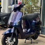 Paarsblauwe retro-scooter op standaard voor een showroom, met ruitpatroon zadel en schijfrem.