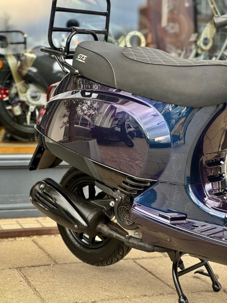 Close-up van de achterzijde van een GTS-scooter met uitlaat en bagagerek, geparkeerd in een showroom.