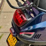 Close-up van de achterkant van een blauw-paarse scooter met bagagerek, achterlicht en gele kentekenplaat.