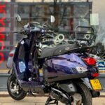 Donkerpaarse GTS-scooter op standaard voor een winkelruit, achteraanzicht met bagagerek en kenteken.