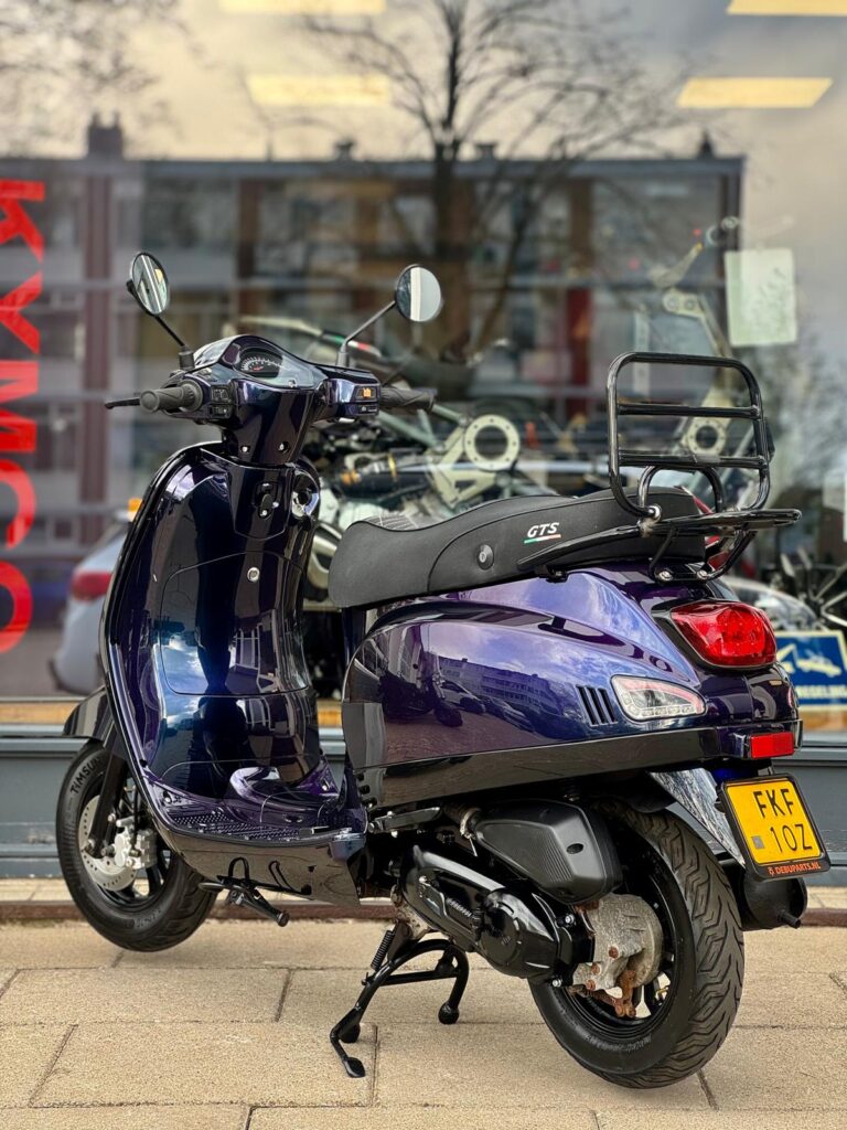 Donkerpaarse GTS-scooter op standaard voor een winkelruit, achteraanzicht met bagagerek en kenteken.