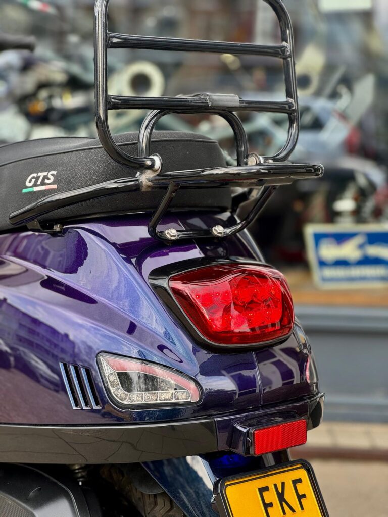 Achterkant van een paarse Vespa GTS-scooter met bagagedrager, achterlicht en knipperlichten.