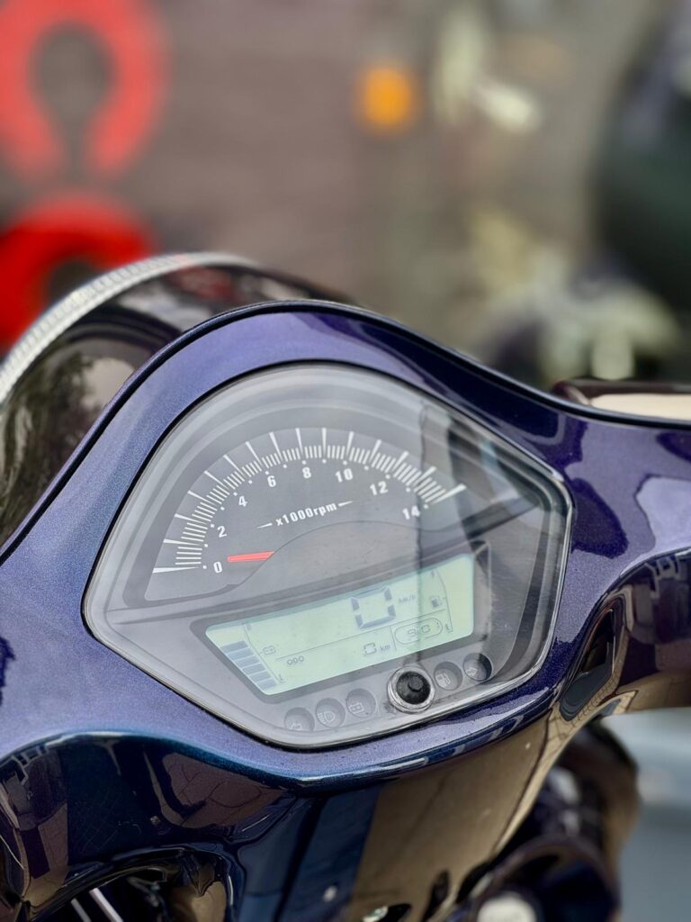 Close-up van een scooter- of motordashboard met toerenteller en digitaal display in een donkerblauwe kuip.