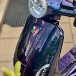 Close-up van een paarse scooter met ronde koplamp, glanzende lak en Italiaans schildembleem op de voorkap.