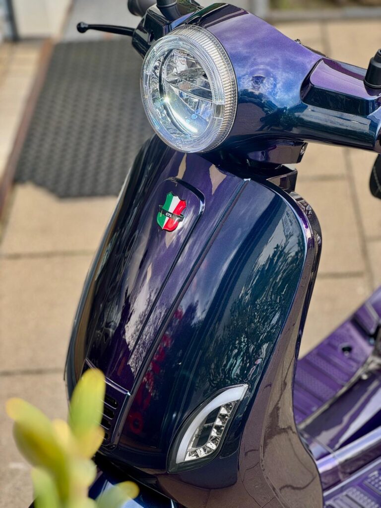 Close-up van een paarse scooter met ronde koplamp, glanzende lak en Italiaans schildembleem op de voorkap.