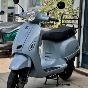 Lichtgrijze retro-scooter met zwart zadel en bagagerek, geparkeerd voor een winkel.