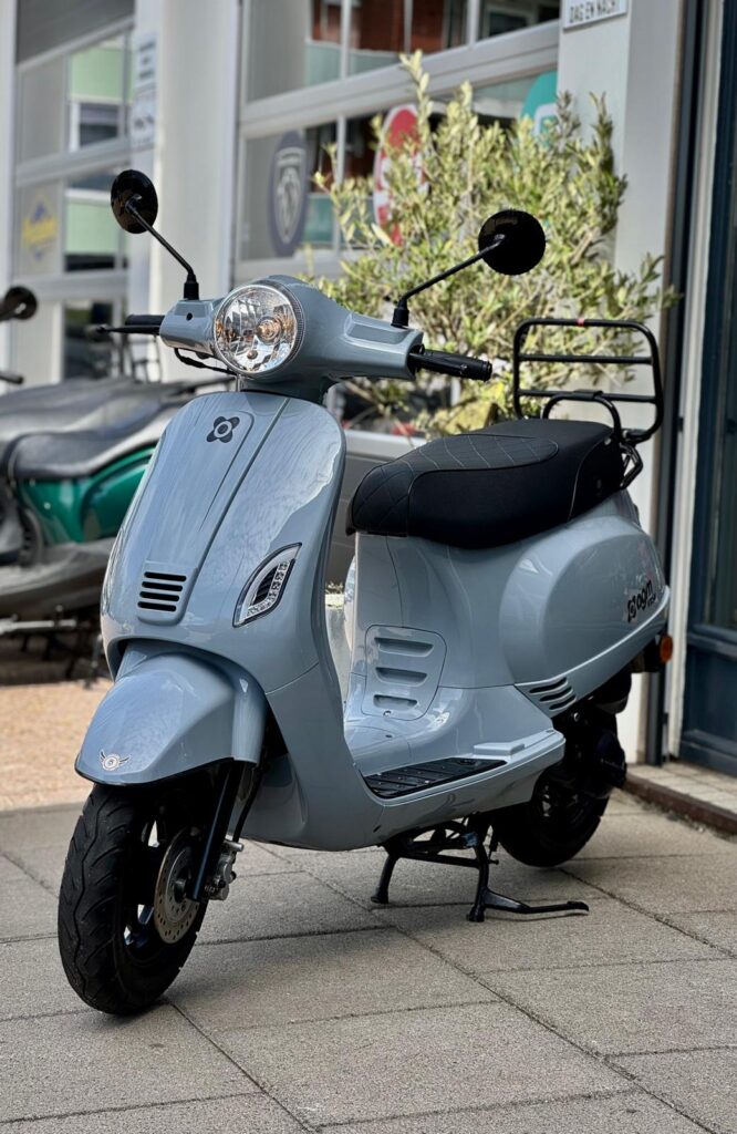 Lichtgrijze retro-scooter met zwart zadel en bagagerek, geparkeerd voor een winkel.