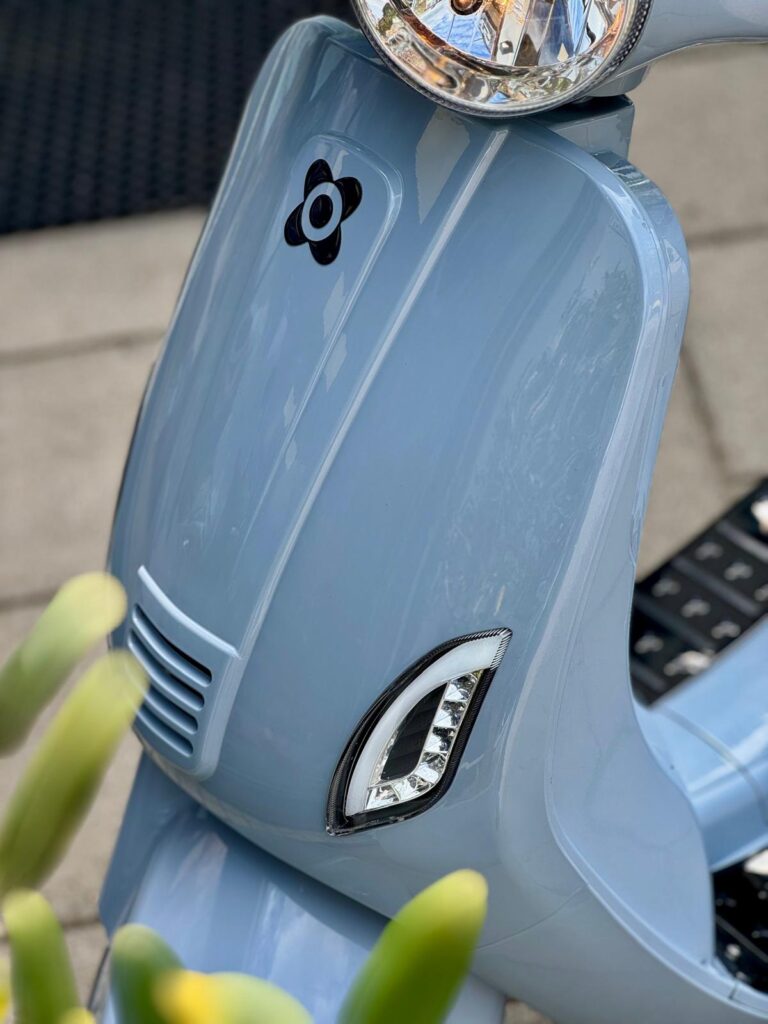 Close-up van de voorkant van een lichtblauwe scooter met koplamp en LED-knipperlicht.