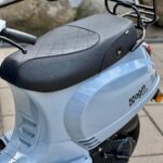 Close-up van lichtblauwe scooter met zwart zadel en bagagedrager op bestrating.