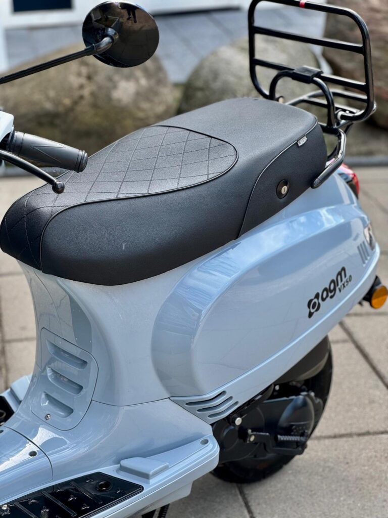 Close-up van lichtblauwe scooter met zwart zadel en bagagedrager op bestrating.