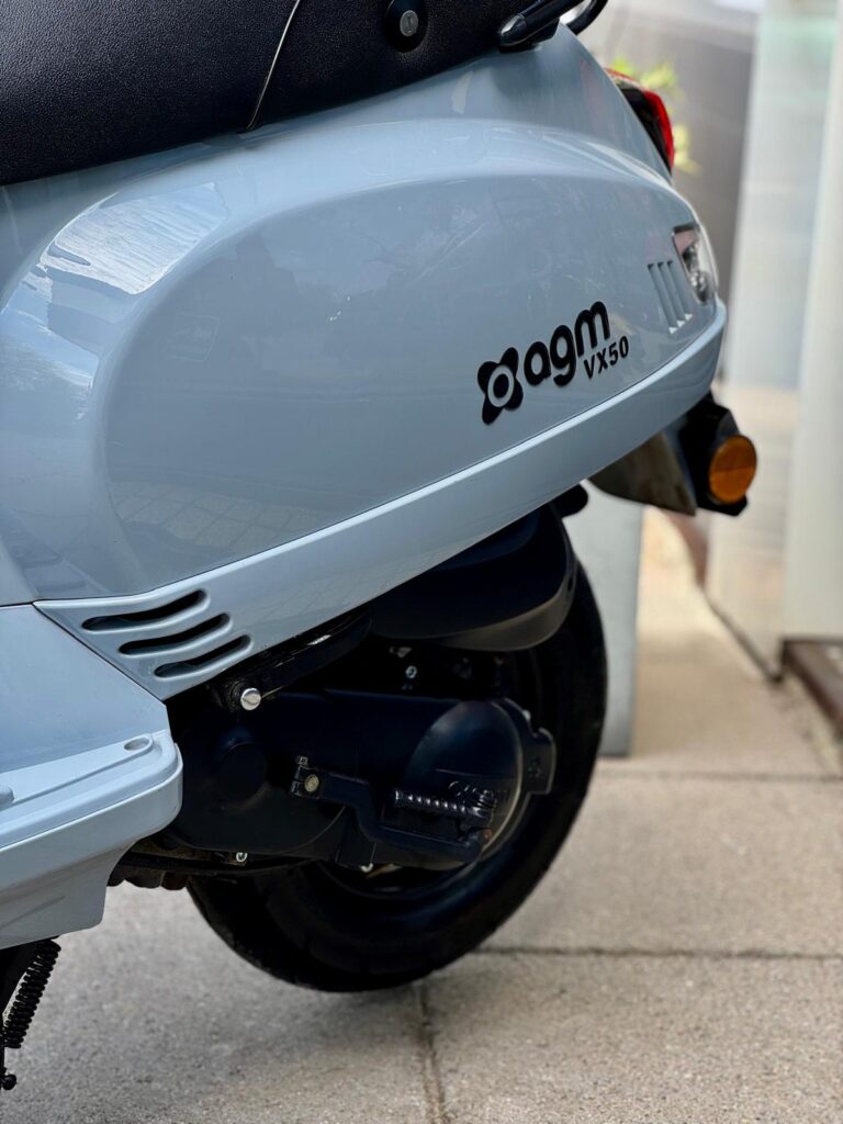 Close-up van de achterzijde van een lichtblauwe agm VX50 scooter met wiel en uitlaat op stoeptegels.