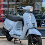 Lichtblauwe retro scooter met zwarte velgen, geparkeerd op de stoep voor een winkelraam.