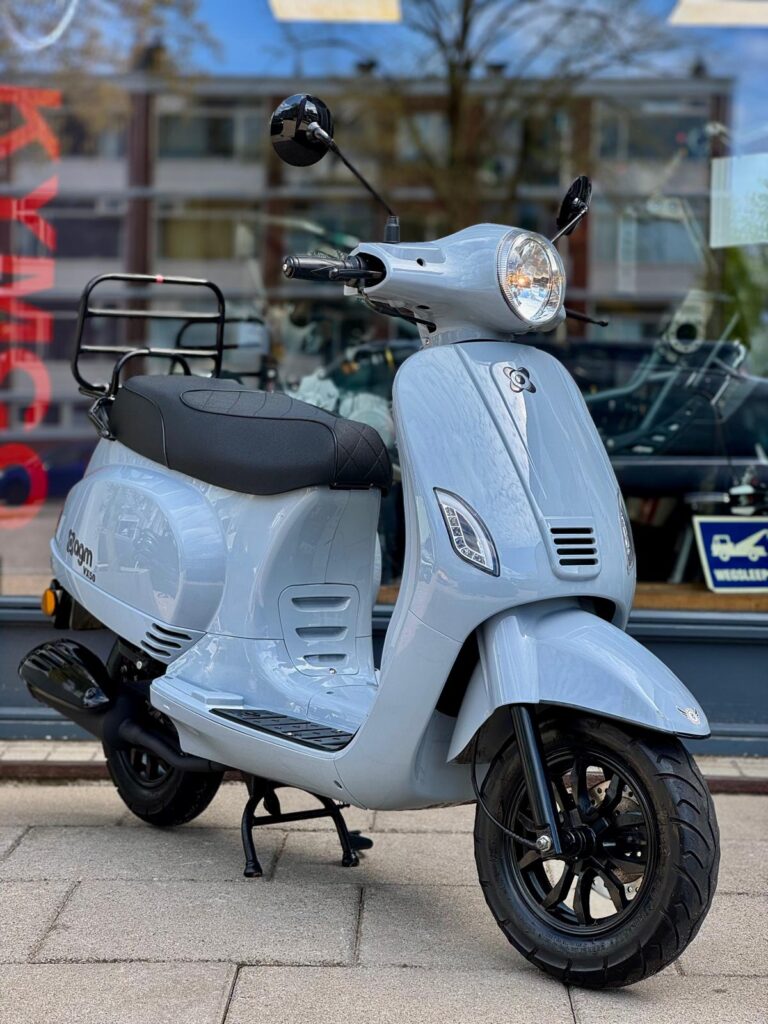 Lichtblauwe retro scooter met zwarte velgen, geparkeerd op de stoep voor een winkelraam.