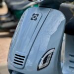 Close-up van de voorkant van een lichtblauwe scooter met ronde koplamp en LED-richtingaanwijzers.