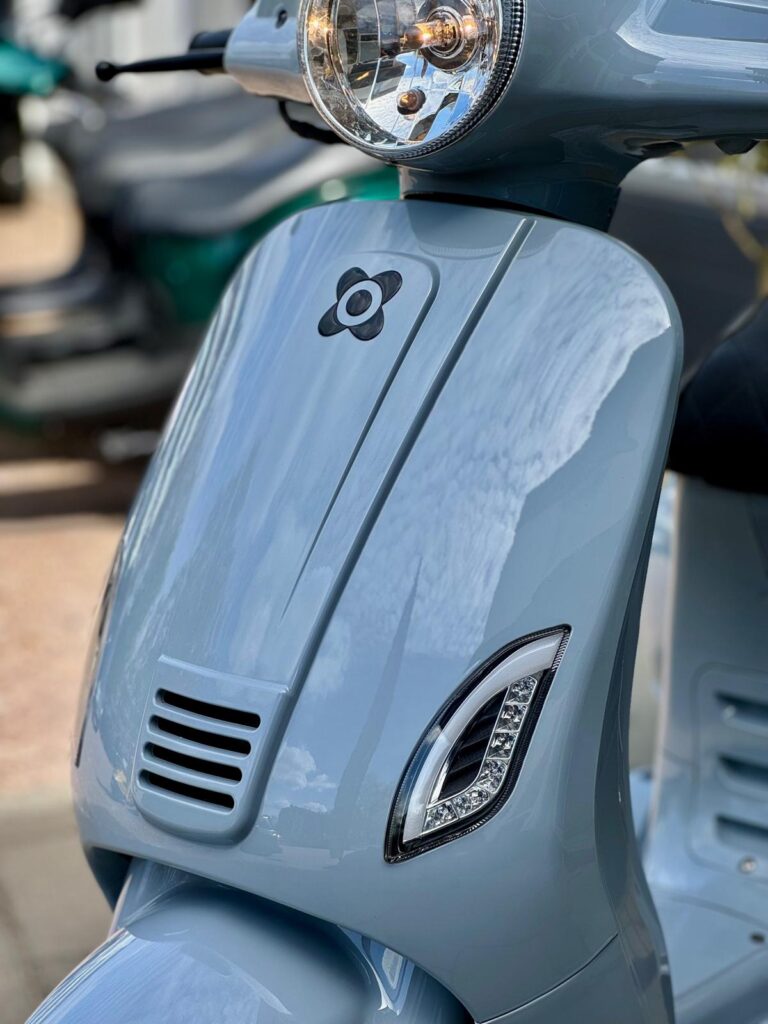 Close-up van de voorkant van een lichtblauwe scooter met ronde koplamp en LED-richtingaanwijzers.