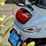 Close-up van de achterkant van een lichtblauwe scooter met bagagedrager, achterlicht en blauwe kentekenplaat.