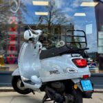 Lichtgrijze AGM VX50 scooter met bagagedrager en blauwe kentekenplaat, geparkeerd voor een winkelpui.
