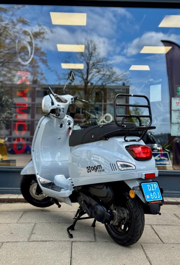 Lichtgrijze AGM VX50 scooter met bagagedrager en blauwe kentekenplaat, geparkeerd voor een winkelpui.