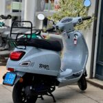 Lichtgrijze AGM VX50 scooter met blauw kenteken, geparkeerd voor een scootershop.