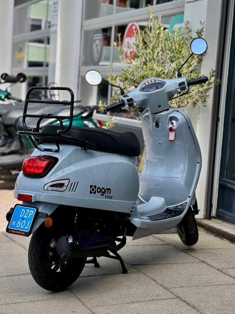 Lichtgrijze AGM VX50 scooter met blauw kenteken, geparkeerd voor een scootershop.