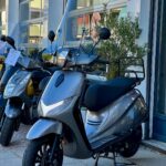 Grijze scooter met hoog windscherm op standaard voor een showroom, met andere scooters ernaast.