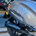 Close-up van het achterwiel en de uitlaat van een grijze Volexim-scooter bij een winkelruit.