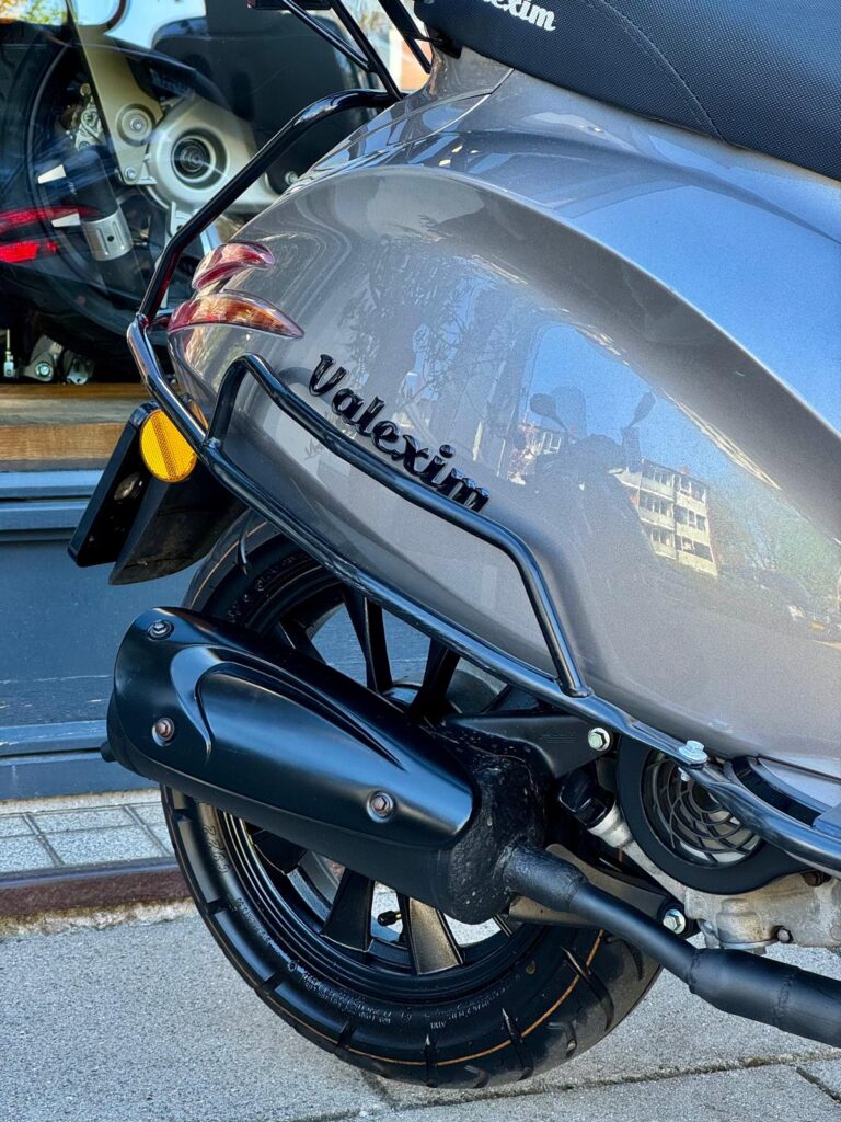 Close-up van het achterwiel en de uitlaat van een grijze Volexim-scooter bij een winkelruit.